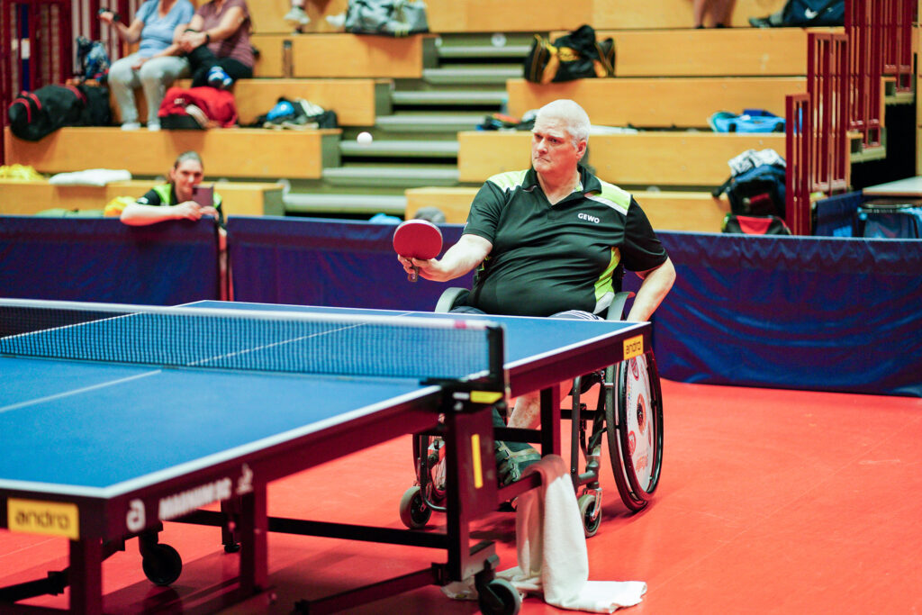 Rollstuhlfahrer spielt Tischtennis in einer Sporthalle.